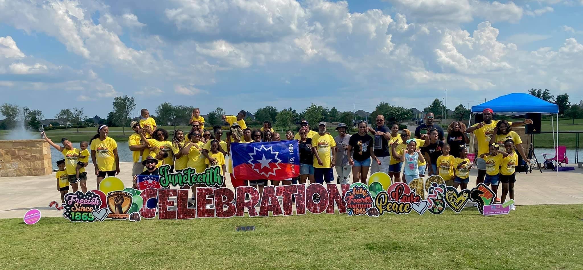 A large group of families in yellow shirts behind a Juneteenth Celebration sign beside a lake